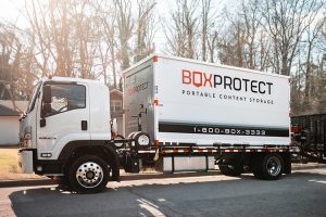 A white BoxProtect portable storage container being delivered by a branded delivery truck in a residential neighborhood. The container features the BoxProtect logo and tagline “Portable Content Storage,” along with the customer service number 1-800-BOX-3333. The image showcases professional mobile storage delivery services, ideal for moving, renovation, and on-site storage solutions.