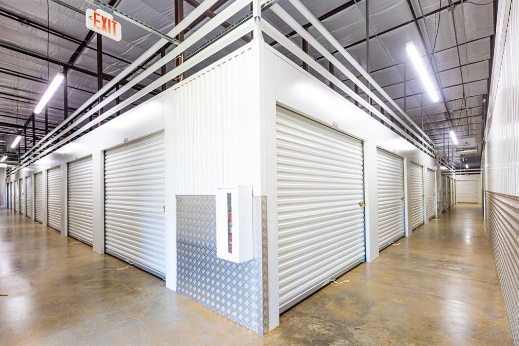 Boxwell indoor self-storage hallway system featuring steel roll-up doors, diamond-plate wainscot panels, integrated burglar bars, and a wall-mounted fire extinguisher, designed for secure, code-compliant interior storage facility construction with clean, durable finishes.